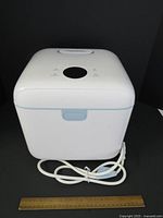 Front angled photo of the Papablic sterilizer showing closed white device with blue latch and attached electric cord, minor surface imperfections noted, ruler in front for scale.