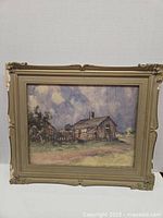 Full view of the painting showing rustic cabin landscape with cloudy sky, in an ornate frame with signs of wear.
