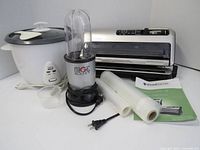 FoodSaver vacuum sealer, Magic Bullet blender with single cup, and rice cooker with lid and rice paddle displayed together on white surface