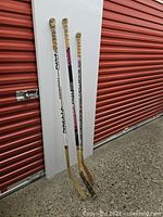 Three wood and composite hockey sticks standing against a white panel and red garage door.