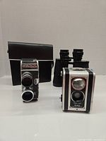 Three vintage camera items together: Bolex movie camera, Kodak Duaflex box camera, and Greif binoculars with leather case in background.