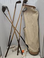 Several vintage golf clubs leaning against a beige antique-style golf bag with golf balls and boxes beside the bag on cloth surface.