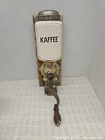 Front view of vintage wall-mounted coffee grinder showing ceramic hopper marked 'KAFFEE' with hand crank and wooden mounting board.
