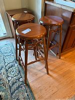 Three dark wood bar stools shown standing on hardwood floor near kitchen cabinetry and a rug. Each stool has a flat round seat and multiple turned wood foot rests between legs.