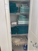 Overview of closet shelf showing neatly folded turquoise and light blue bath towels on three shelves and two cream-colored bathroom rugs on the bottom shelf.