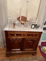 Full frontal view of the wooden cabinet showing the marble top with various decorative objects on top. The cabinet has two doors and a drawer with carved wooden details.