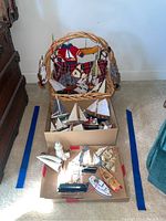 Lot overview showing wicker basket with fabric sailboat ornaments and two boxes with wooden sailboats, lighthouse, and other Christmas nautical-themed ornaments.