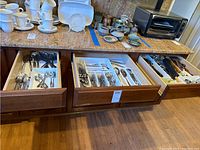 Photo showing three kitchen drawers with various flatware and cooking utensils inside, plus some kitchenware on countertop.