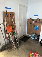 Full view of gardening area with large tools like shovels, rakes, and a snow shovel leaning against a pegboard on the left side, with the door in background.