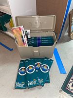 Open plastic storage case showing multiple unopened packs of oil pastels and a set of colored pencils