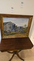 Framed oil painting showing farm and barn with rustic wooden frame on a wooden table.