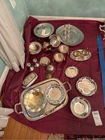 Wide view showing all silverplate items arranged on a maroon fabric including trays, bowls, condiment holders, salt and pepper shakers, and candlesticks.