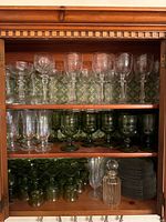 Wooden cabinet with three shelves holding various glasses and plates: top shelf with clear etched wine glasses, middle shelf with green wine and water glasses and champagne flutes, bottom shelf with more green glasses, clear decanter, and black plates.
