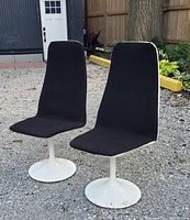 Front view of two 1960s chairs showing black fabric seating surfaces and white painted round metal bases with some rust and paint wear.