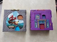 Front view of two closed fold-out playset cases, one featuring a pirate ship illustration in a gray rock-like frame and the other a purple spooky mansion with door and window details.