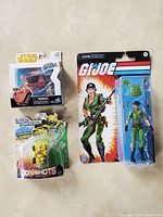 Three sealed items displayed: a Star Wars helmet figure in white box with clear window, a Transformers BotShots Bumblebee figure, and a G.I. Joe Lady Jaye action figure with accessories in blister pack.
