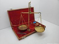 Antique miniature balance scales set displayed in open wooden case with two brass weighing pans suspended from balance beam, red velvet lining visible inside the case.