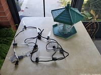 Photo of green birdfeeder with hexagonal roof and base on a table alongside black wire string lights with clear tubular bulbs