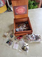 Open wooden Kristoff cigar box with multiple pins inside and several sealed plastic bags of pins arranged beside it on a tabletop.