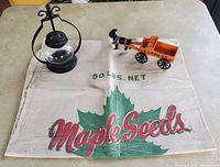 Maple Seeds cloth seed sack, cast iron cart labeled 'EXPRESSO', and mini hanging oil lantern on beige table.