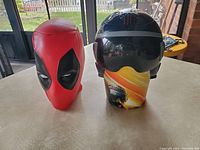 Two items on a table: red Deadpool candy head and black Top Gun Maverick pilot helmet cup