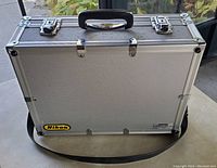 Front and side view of the closed aluminum camera case showing Nikon logo, handle, dual locking latches, and shoulder strap.