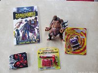 Full view of the lot showing the comic, monster figure, Deadpool wallet, rubber band gun in packaging, and wristband dispenser in packaging on a beige surface.