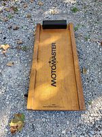 Top view of wooden mechanic creeper board placed on gravel, showing MotorMaster logo and black padded headrest.