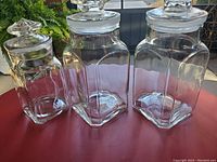 Three vintage heavy glass square canister jars with faceted glass stoppers displayed on red surface outdoors. No cracks, some knicks on stoppers.