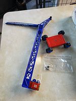 Photo showing the blue lattice tin crane with a red wheeled base and a string hook, laid out on a table