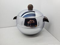 Front and side view of the vintage West Bend penguin hot and cold server showing reflective chrome body, wooden handles, and embossed penguin motifs.