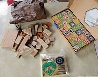 Photo showing wooden building blocks of various shapes spread out with a brown bag, vintage alphabet blocks in original box, and a sealed vintage baseball peg game package on tabletop.