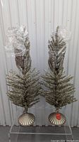 Two gold tinsel Christmas trees standing side by side on clear shelving, topped with plastic wrapping to protect the tips.