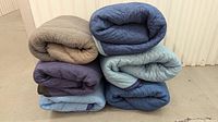 Stack of six folded moving blankets in various shades of blue, purple, and gray, showing texture and fabric detail.