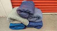 Five folded and stacked moving blankets in different blue and gray colors, placed next to a red storage unit door, showing colors and condition.