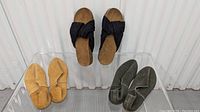 Front view of three pairs of suede women's shoes: black sandals (size 7), mustard yellow slip-ons (size 6), olive green slip-ons (size 6) on clear display stand.
