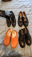 Four pairs of shoes on bed: black leather boots, black lace-up espadrilles, orange clogs, black dress shoes.