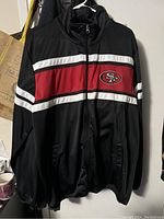 Front view of the black San Francisco 49ers men's track jacket with red and white stripes and 49ers logo.