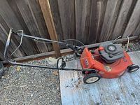 Side view of red gas powered push lawnmower on gravel and metal sheet, showing handle, wheels and engine cover.