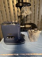 Side view of the Orfeld electric juicer showing the dark blue main body and black attachments wrapped in plastic.