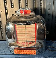 Front view of chrome retro jukebox style radio with detailed selection letters and numbers at bottom, and red and white song or station list visible behind a glass panel.