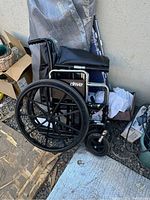 Manual wheelchair branded Drive Medical with black seat and backrest. Large rear wheels and smaller front caster wheels visible, sitting outside with miscellaneous items around.
