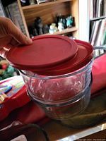 Photo of two clear Pyrex glass bowls stacked, each with a red lid being held by a hand.
