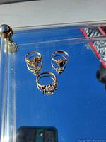 Photo shows three rings on a reflective surface with varied gemstone settings and metal types.