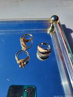 Three rings displayed on a reflective surface under sunlight, showing detailed designs and stone embellishments.