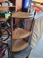 Three-tier wooden corner shelf with woven cane surfaces, displaying turned spindle supports and finials on top.