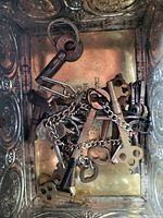 Photo showing a variety of vintage metal keys and chains in a metallic container with embossed pattern.