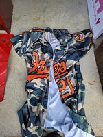 Front view of camouflage baseball jersey laid out, showing orange 'Orioles' script and number '21' with Baltimore Orioles logo patch on sleeve.