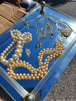 Photo showing assorted jewelry items including faux pearl necklaces, earrings, pendant necklace, and a brooch or ring on a mirrored glass tray