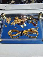 Photo of assorted gold-tone jewelry laid out on a reflective surface, including a thick mesh bracelet, chain necklaces, clip-on earrings, and pins.
